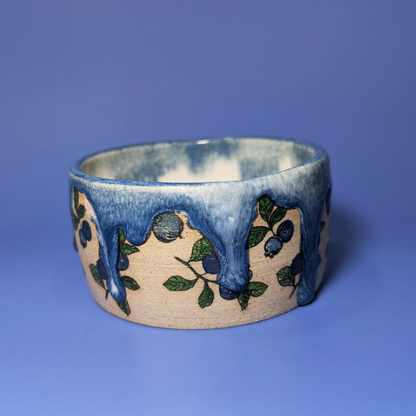 A handmade ceramic wide bowl featuring a cream-colored base decorated with blueberry illustrations and a glossy blue dripping glaze, set against a solid blue background.