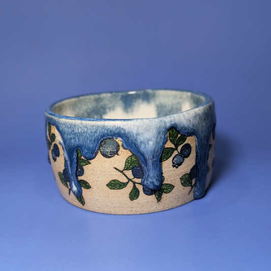 A handmade ceramic wide bowl featuring a cream-colored base decorated with blueberry illustrations and a glossy blue dripping glaze, set against a solid blue background.