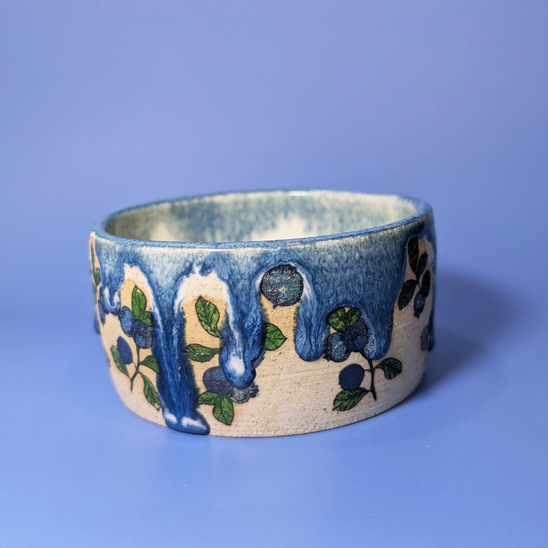 A handmade ceramic wide  bowl featuring a cream-colored base with a blue dripping glaze and blueberry motifs, set against a solid blue background.
