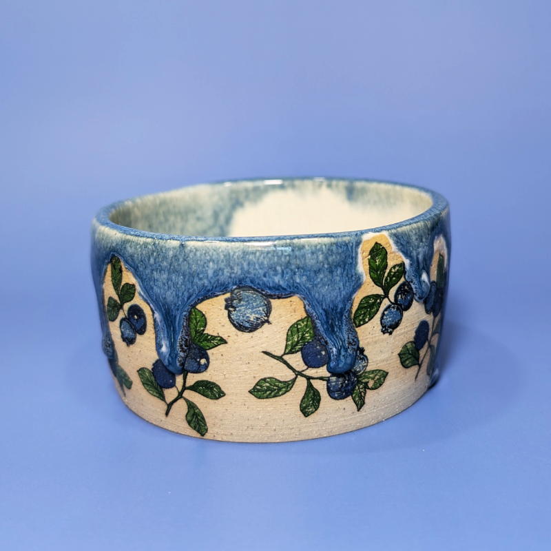 A handmade ceramic bowl featuring a blue drippy glaze design with blueberries and branches, set against a solid blue background.
