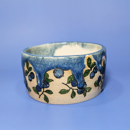 A handmade ceramic bowl featuring a blue drippy glaze design with blueberries and branches, set against a solid blue background.