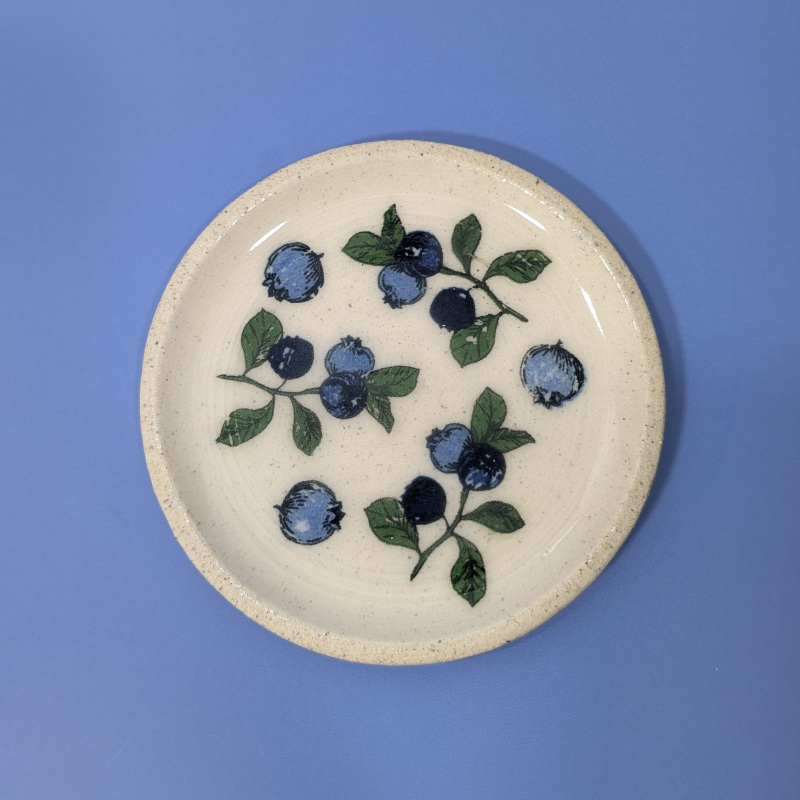 A top down view of this cream colored clay trinket dish. It shows an arrangement of blueberries and leaves.