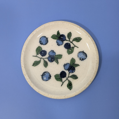 A top down view of this cream colored clay trinket dish. It shows an arrangement of blueberries and leaves.