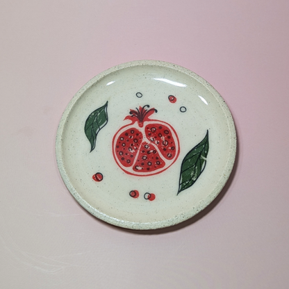 A trinket dish showing a red-orange pomegranate sliced in half. Green leaves  and small seeds encircle the fruit.